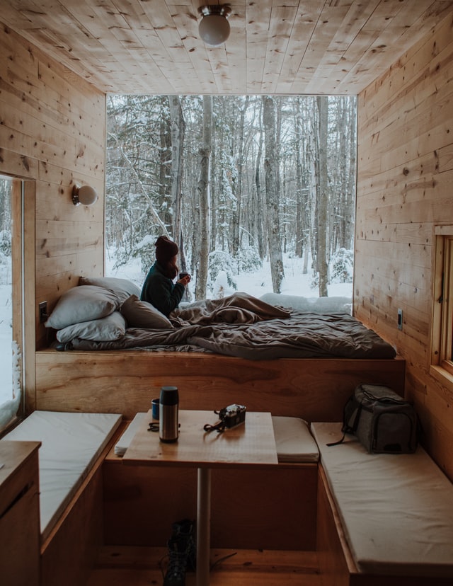 Ein minimalistisch eingerichtetes Innere eines Tiny House ist zu sehen: Es ist komplett mit Holz verkleidet und an der Fensterfront ist ein Bett, in dem eine Frau sitzt, die aus dem Fenster schaut. 