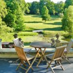 Auf einer mit grauen Steinplatten ausgelegten Terrasse mit blick auf einen grünen Park stehen Gartentisch und Stühle