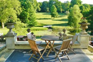 Auf einer mit grauen Steinplatten ausgelegten Terrasse mit blick auf einen grünen Park stehen Gartentisch und Stühle