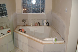 Eine Badsanierung im bewohnten Zustand: Eine Eckbadewanne, noch nicht komplett eingefliest, nur mit Gipskartonplatten verkleidet, auf deren Rand verschiedene Dusch- und Badeartikel stehen.