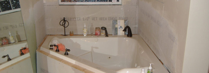 Eine Badsanierung im bewohnten Zustand: Eine Eckbadewanne, noch nicht komplett eingefliest, nur mit Gipskartonplatten verkleidet, auf deren Rand verschiedene Dusch- und Badeartikel stehen.