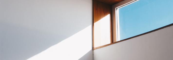 Eine Ecke im Inneren eines Hauses ist zu sehen. Die Wände sind weiß und glatt, oben ist ein Teil eines waagerechten, rechteckigen Fensters zu sehen, durch das die Sonne hereinscheint.