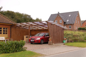 Ein Haus in einer Wohngegend, an dessen Seite ein Carport aus Holz mit durchsichtigem Dach gebaut wurde. Darin steht ein rotes Auto.