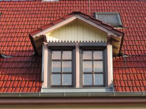Eine Dachgaube aus hellem Holz und mit zwei Fenstern ragt mittig aus einem mit roten Dachziegeln gedecktem Dach heraus.