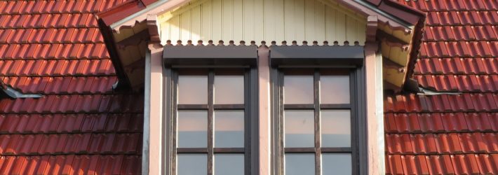 Eine Dachgaube aus hellem Holz und mit zwei Fenstern ragt mittig aus einem mit roten Dachziegeln gedecktem Dach heraus.