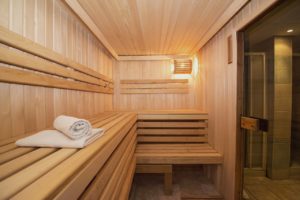 Eine große Sauna von innen. Sie ist mit hellem Holz vertäfelt und hat Bänke aus Holz. Rechts ist der Eingang, links liegen weiße Handtücher auf den Bänken.
