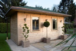 Eine Außensauna aus Holz. Das kleine Häuschen aus hellem Holz steht auf einem Untergrund aus schönen Steinplatten in einem Garten.