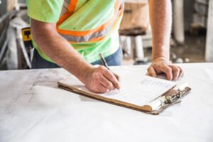Ein Bauarbeiter füllt ein Formular aus, was auf einem Tisch liegt, auf dem verschiedene Pläne ausgebreitet sind.