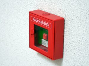 An einer weißen Wand hängt ein roter Rauch- bzw. Brandmelder.