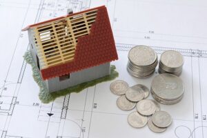 Auf einigen Papieren und Plänen steht ein kleines Haus-Modell mit halb fertigem Dach, daneben liegen einige Münzen.