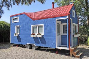 Ein Tiny House auf Rädern ist zu sehen. Es hat eine blaue Holzverkleidung und ein rotes Dach.