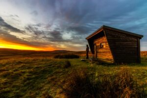 Ein Tiny House aus Holz steht mitten in einer einsamen Landschaft, davor steht eine Person und schaut in den Sonnenuntergang