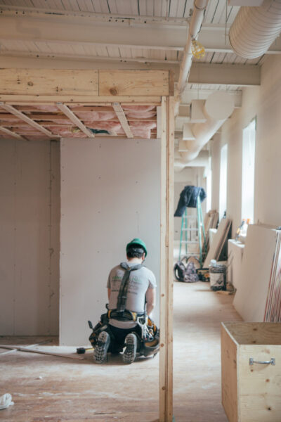 Ein Mann kniet auf einem Boden aus Holzfasern. Um ihn herum wird ein neues Haus gebaut